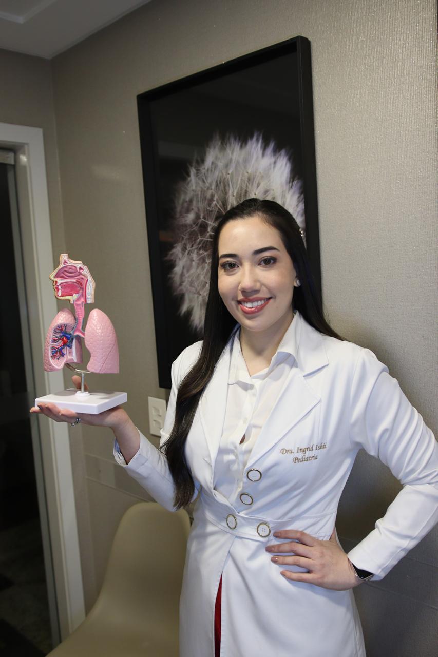 Dra. Ingrid Ishii de jaleco, segurando um modelo do sistema respiratório no consultório